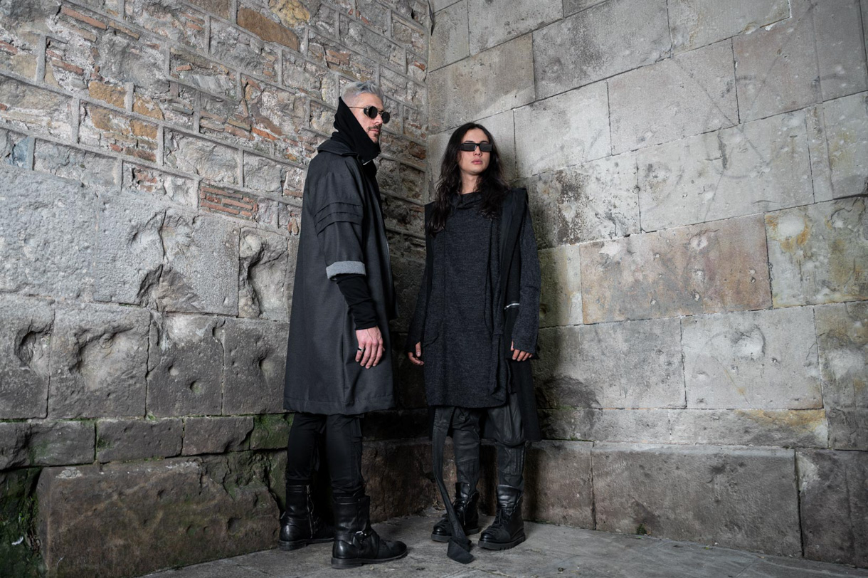 Two models dressed in black beside a stone wall