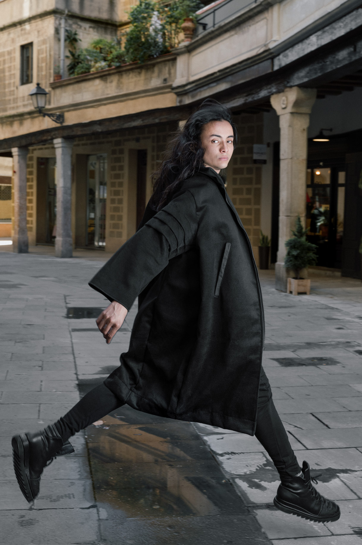 Model in a long black coat walking through an arcaded square