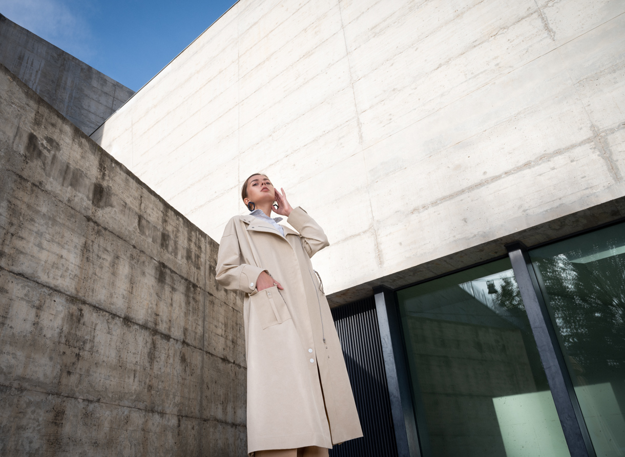 Model in a beige coat photographed from below beside a concrete wall