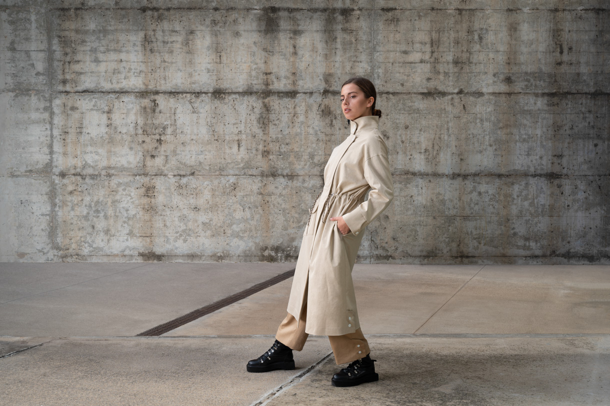 Model in a beige trench coat walking in front of a concrete wall