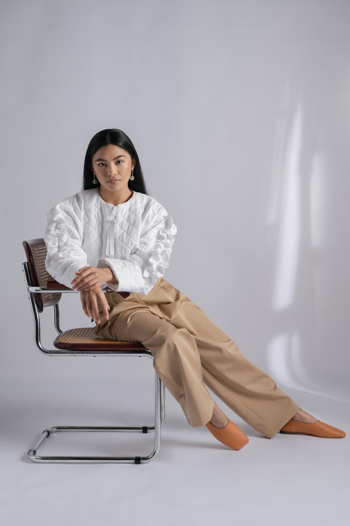 Model seated on a chair in a white blouse and camel trousers