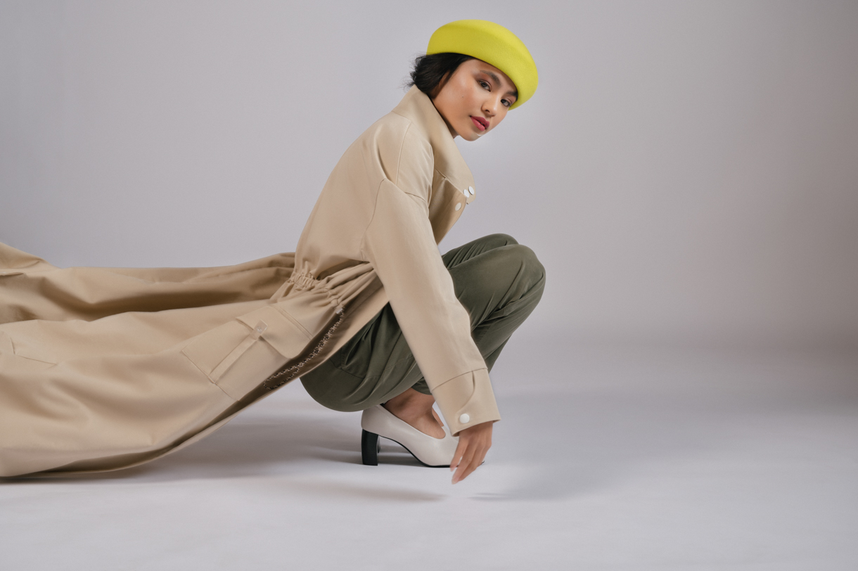 Model crouching in a beige trench coat and yellow hat in the studio